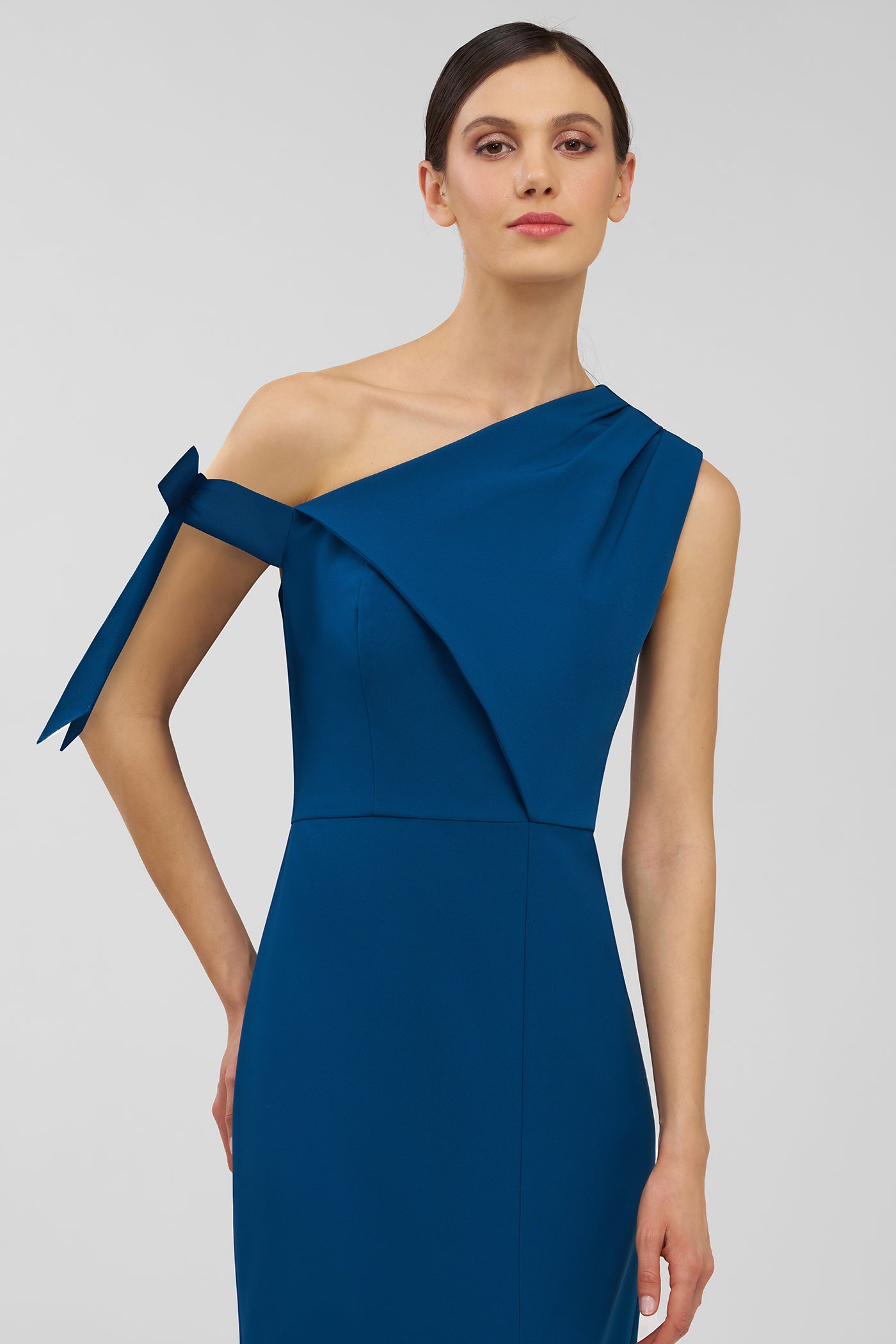 Woman wearing a blue one-shoulder dress with a bow detail on a light gray background