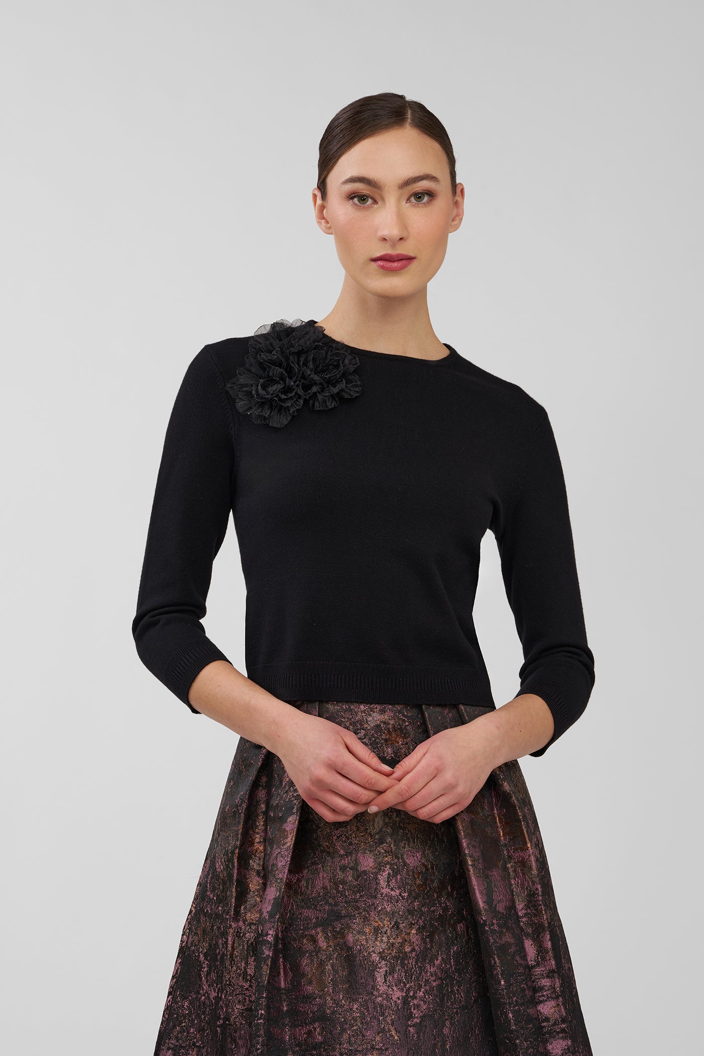 Woman wearing a black top with a floral detail and a patterned skirt on a plain background