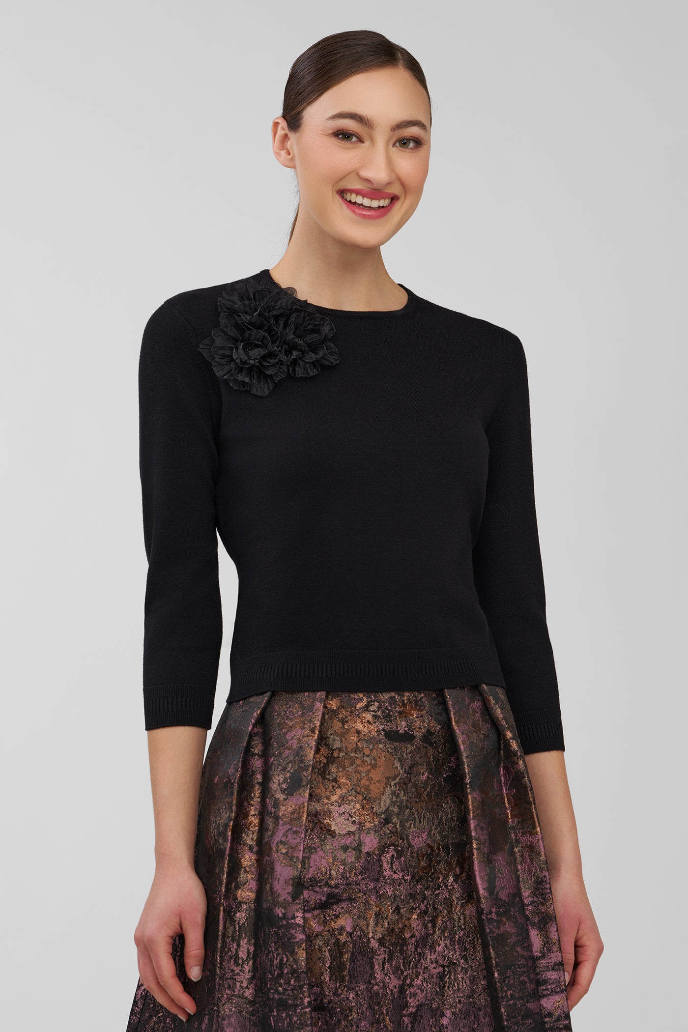 Woman wearing a black long-sleeve top with a floral detail and a patterned skirt on a plain background
