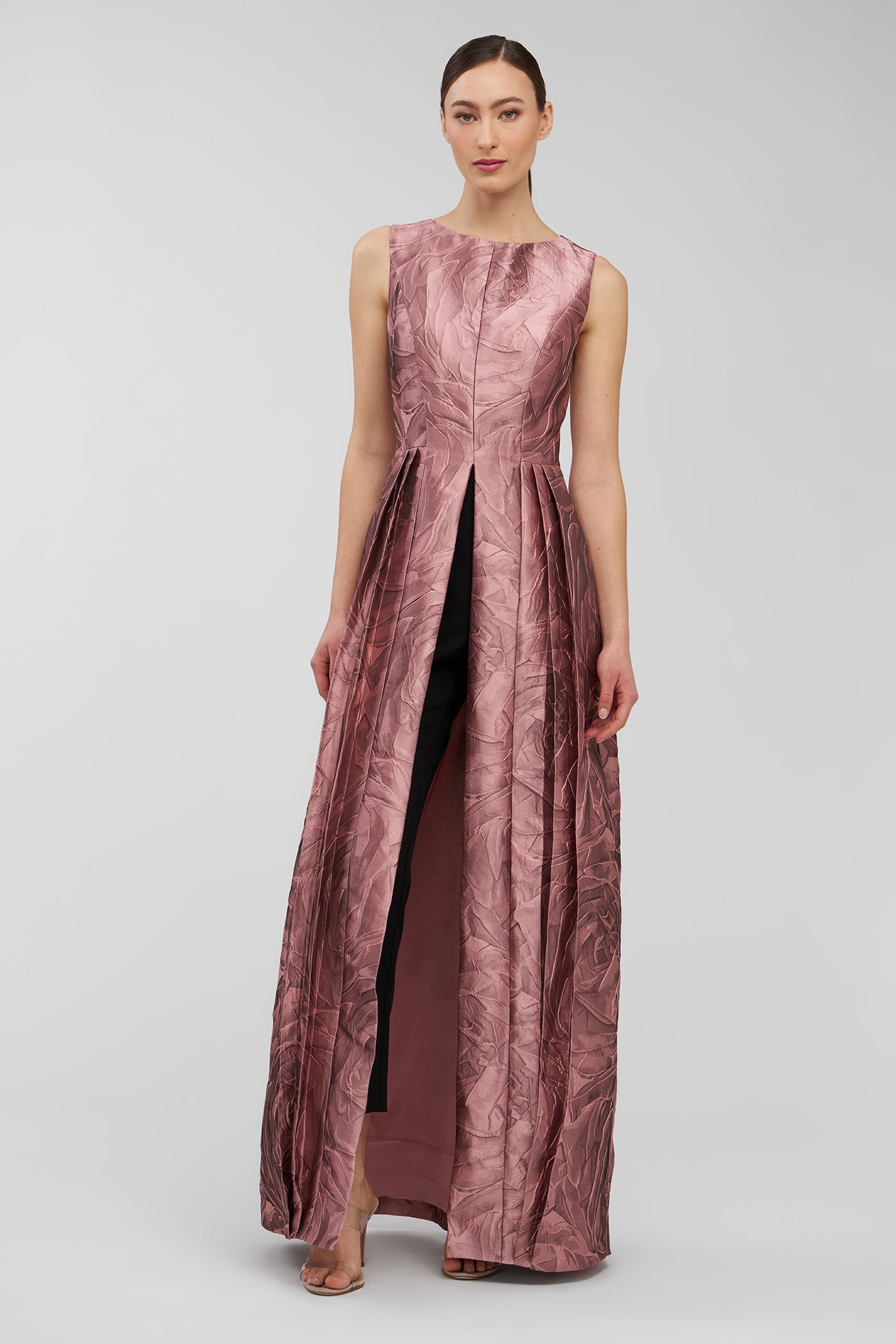 Woman wearing a pink evening walk thru jumpsuit with a slit on a light gray background