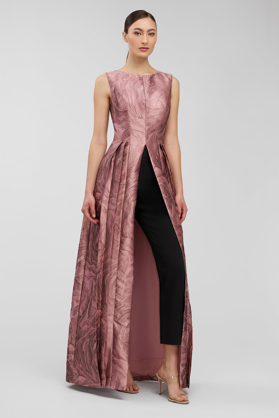 Woman wearing a long, pleated, pink walk thru jumpsuit with a fitted brown trousers underneath on a white background