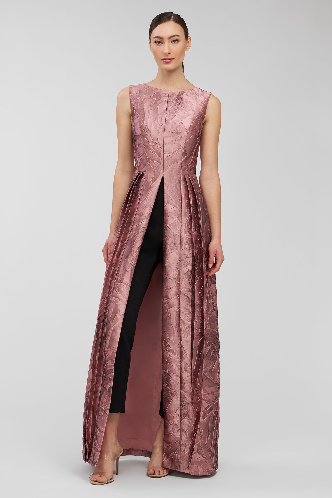 Woman wearing a long, sleeveless, metallic pink walk thru jumpsuit on a light gray background